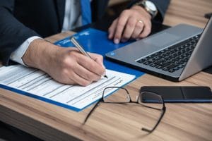 Person filling papers with a laptop and phone in the background
